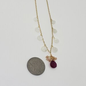 Op de afbeelding: Een gouden ketting met een cluster van kleine roze en rode edelstenen die aan de onderkant hangen. De ketting is versierd met verschillende witte maansteenbedels.