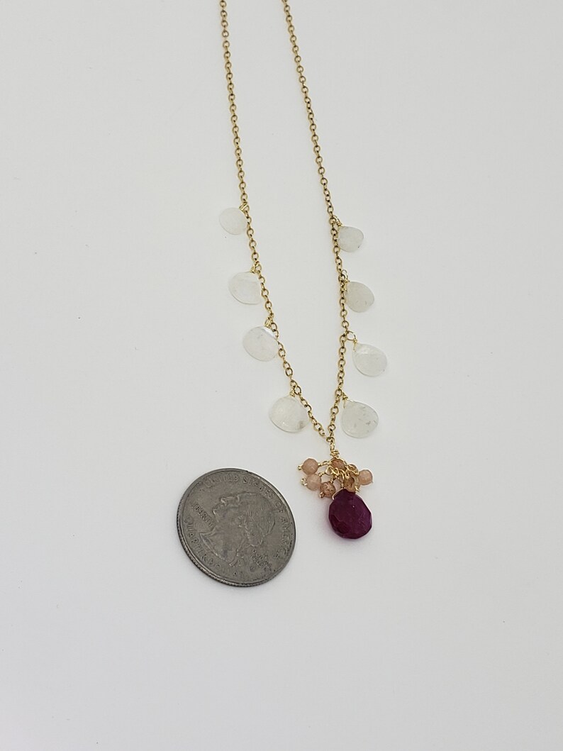 Op de afbeelding: Een gouden ketting met een cluster van kleine roze en rode edelstenen die aan de onderkant hangen. De ketting is versierd met verschillende witte maansteenbedels.