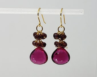May include: A pair of gold-colored drop earrings. Each earring features a cluster of small, dark red beads above a large, faceted, teardrop-shaped, magenta-colored stone. The earrings hang from gold-colored hooks.