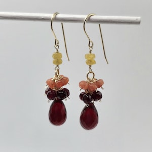 May include: A pair of gold-tone drop earrings featuring a yellow faceted bead at the top, followed by a cluster of small peach-colored beads, a layer of dark red faceted beads, and a large, teardrop-shaped dark red faceted gemstone at the base.