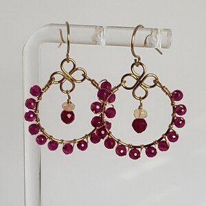 May include: Gold-toned hoop earrings with a floral design at the top and a cluster of small, round, deep pink gemstones around the hoop. Each earring has a single, larger pink gemstone and a small, oval, off-white gemstone dangling from the center.