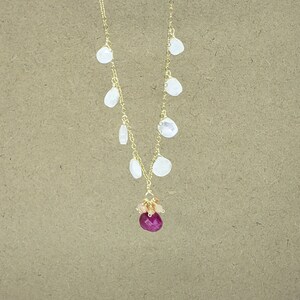 Op de afbeelding: Een gouden ketting met een cluster van roze en oranje edelstenen en een grote, gefacetteerde, robijnrode edelsteenhanger. De ketting heeft ook een reeks kleinere, witte, druppelvormige edelstenen die aan de ketting hangen.