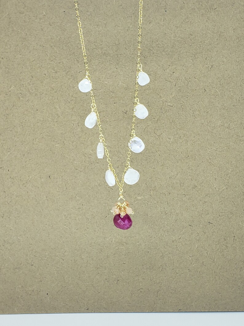 Op de afbeelding: Een gouden ketting met een cluster van roze en oranje edelstenen en een grote, gefacetteerde, robijnrode edelsteenhanger. De ketting heeft ook een reeks kleinere, witte, druppelvormige edelstenen die aan de ketting hangen.