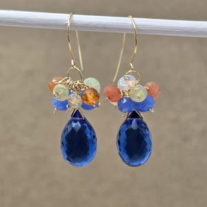 May include: A pair of gold-tone earrings featuring a cluster of small orange, green, and blue beads above a large, faceted, teardrop-shaped blue gemstone. The earrings are suspended from gold-colored ear wires.