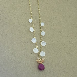 Op de afbeelding: Een gouden ketting met een cluster van kleine, witte maansteenstenen en een grotere, dieprode robijn-steen aan de onderkant.