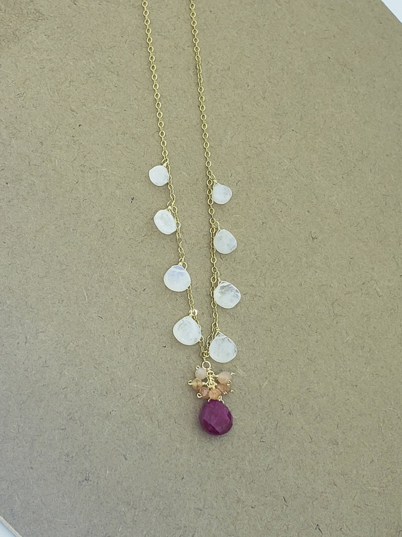 Op de afbeelding: Een gouden ketting met een cluster van kleine, witte maansteenstenen en een grotere, dieprode robijn-steen aan de onderkant.