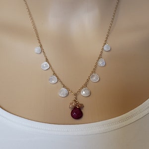 Op de afbeelding: Een gouden ketting met een rode edelsteen hanger en witte maansteen accenten. De ketting heeft een cluster van kleinere edelstenen aan de onderkant van de hanger.