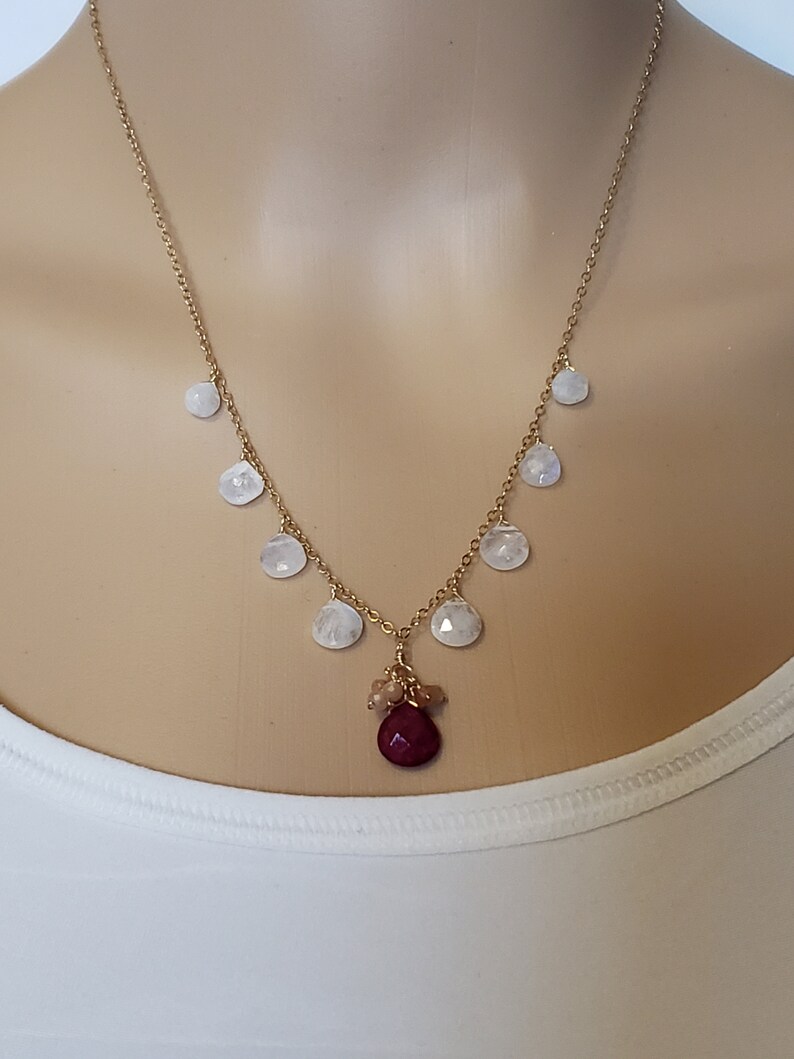 Op de afbeelding: Een gouden ketting met een rode edelsteen hanger en witte maansteen accenten. De ketting heeft een cluster van kleinere edelstenen aan de onderkant van de hanger.