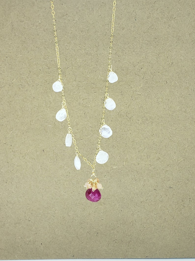 Op de afbeelding: Een gouden ketting met een roze edelsteen hanger en witte edelsteen accenten. De ketting heeft een cluster van kleinere roze edelstenen die de grotere roze edelsteen omringen.