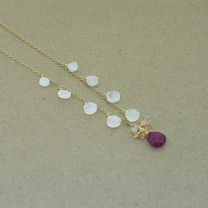 Op de afbeelding: Een gouden ketting met een cluster van kleine, lichtroze en oranje stenen en een grotere, donkerroze steen aan de onderkant. De ketting is versierd met verschillende witte, ovale stenen.
