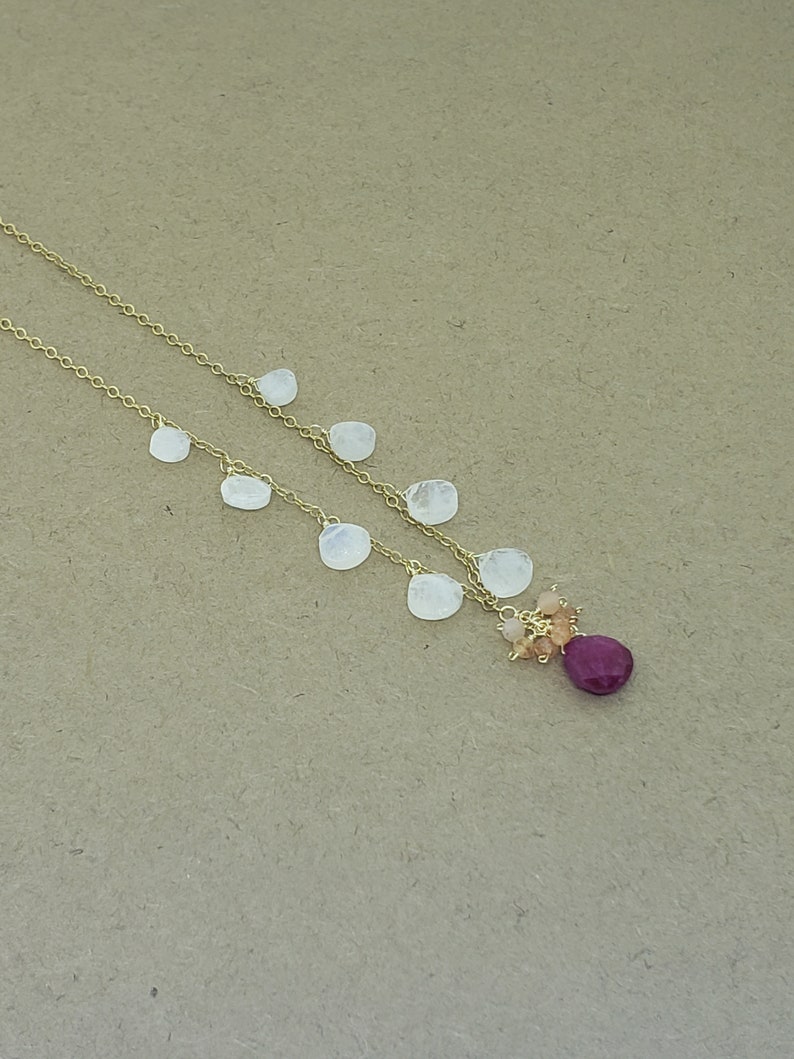 Op de afbeelding: Een gouden ketting met een cluster van kleine, lichtroze en oranje stenen en een grotere, donkerroze steen aan de onderkant. De ketting is versierd met verschillende witte, ovale stenen.