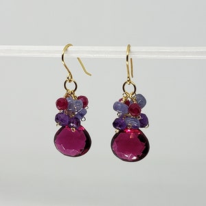 May include: A pair of gold-colored drop earrings. Each earring features a large, faceted, teardrop-shaped, magenta-colored gemstone at the bottom. Above the magenta stone are clusters of smaller purple, red, and blue gemstones.