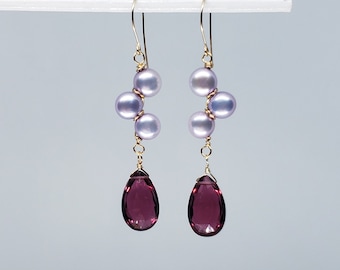 May include: A pair of gold earrings with a teardrop-shaped purple gemstone dangling from each earring. Three small, light purple pearls are clustered above each gemstone.