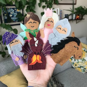May include: A collection of felt finger puppets featuring a fairy, princess, knight, wizard, dragon, and horse. The puppets are in various colors, including purple, pink, brown, and green, with gold and silver accents. The puppets are designed with smiling faces.