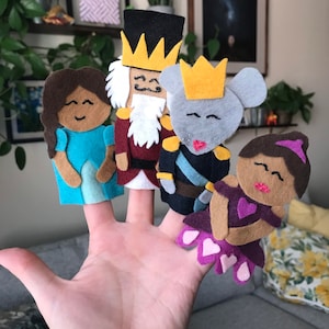 May include: A hand displaying five felt finger puppets. The puppets include a girl in a blue dress, a nutcracker, a mouse king, and a ballerina. The puppets are made of felt in various colors, including blue, brown, gray, red, and purple.