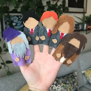 Op de afbeelding: Een hand die vijf vingerpoppetjes van vilt toont. De poppetjes zijn onder meer een tovenaar in paars, drie figuren in grijze pakken met rode stropdassen en een bebaarde figuur met bruin haar. De poppetjes zijn ontworpen met eenvoudige gelaatstrekken.