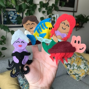 Op de afbeelding: Een verzameling vingerpoppetjes van vilt met personages uit een populaire animatiefilm. De poppetjes omvatten een zeemeermin met rood haar, een geel-blauwe vis, een paarse zeeheks, een man met bruin haar en een roze krab.