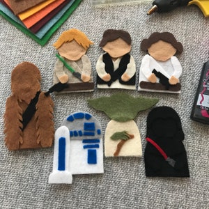 May include: Handmade felt Star Wars character figures, including Luke Skywalker, Han Solo, Princess Leia, Chewbacca, R2-D2, Yoda, and Darth Vader. The figures are made of various colors of felt and are arranged on a gray surface.