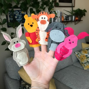 May include: A hand with five felt finger puppets: a grey rabbit, Winnie the Pooh, Tigger, Eeyore, and Piglet. The puppets are colourful, with details like pink ears, red shirts, and orange stripes. The background includes plants and a grey sofa.