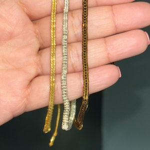 Peut inclure: Trois chaînes de perles de couleur or, argent et marron. Les chaînes sont composées de petites perles carrées et pendent d'une main.