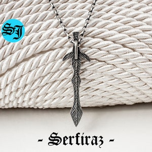May include: A silver sword pendant necklace on a silver chain. The pendant is detailed with a textured design and a pointed tip. The necklace is displayed on a white rope background. -Sertiraz-