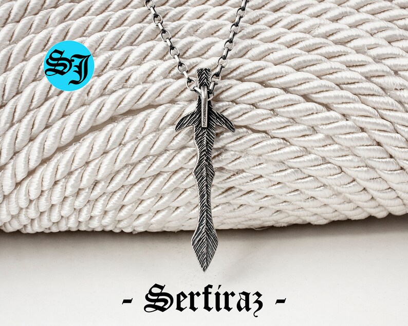 May include: A silver sword pendant necklace on a silver chain. The pendant is detailed with a textured design and a pointed tip. The necklace is displayed on a white rope background. -Sertiraz-