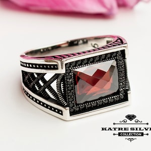 Peut inclure: Une bague en argent avec une pierre rouge entourée de pierres noires. La bague présente un design détaillé avec un motif en forme de croix.