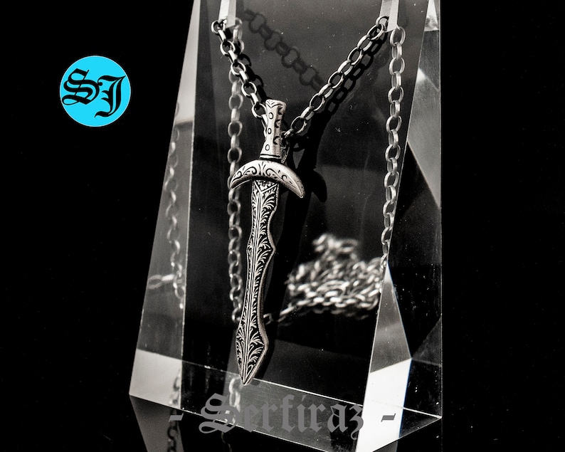 May include: A silver sword pendant necklace with an intricate design. The pendant is hanging from a silver chain.