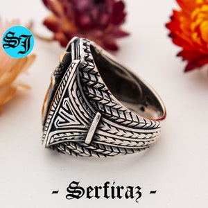 May include: A silver ring with intricate geometric patterns and a gold-colored central design. The ring features a textured band and a triangular centerpiece. The word "- Serfiraz -" is printed at the bottom of the image.