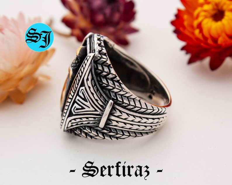 May include: A silver ring with intricate geometric patterns and a gold-colored central design. The ring features a textured band and a triangular centerpiece. The word "- Serfiraz -" is printed at the bottom of the image.