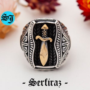 May include: A silver ring with a gold-colored dagger design. The ring has intricate detailing and the text "- Serfiraz -" in a decorative font. The ring is set against a blurred background with flowers.