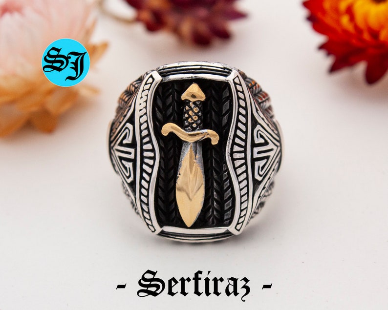 May include: A silver ring with a gold-colored dagger design. The ring has intricate detailing and the text "- Serfiraz -" in a decorative font. The ring is set against a blurred background with flowers.