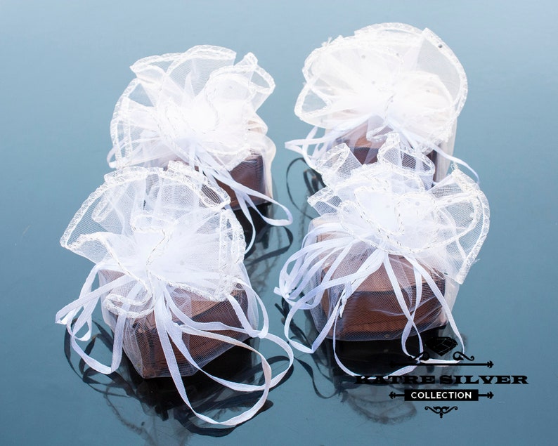 May include: Four small, brown square boxes are wrapped in sheer white fabric and tied with white ribbons. The boxes are arranged on a reflective surface, with the white fabric creating a delicate, airy appearance. The boxes are likely for gifts.
