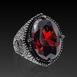 May include: A silver ring with an ornate design and a large, oval-shaped red gemstone.