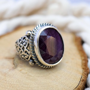 Könnte beinhalten: Ein Silberring mit einem großen, ovalen, tiefvioletten Edelstein. Der Ring hat ein verziertes, durchbrochenes Design am Band und einen gedrehten, seilartigen Rand um den Edelstein. Der Ring wird auf einer Holzoberfläche präsentiert.