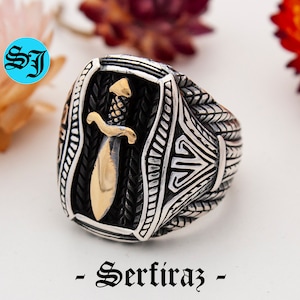 May include: A silver-toned ring with a gold-colored dagger design. The ring has intricate detailing and the text "- Serfiraz -" is at the bottom. The ring is on a white surface.