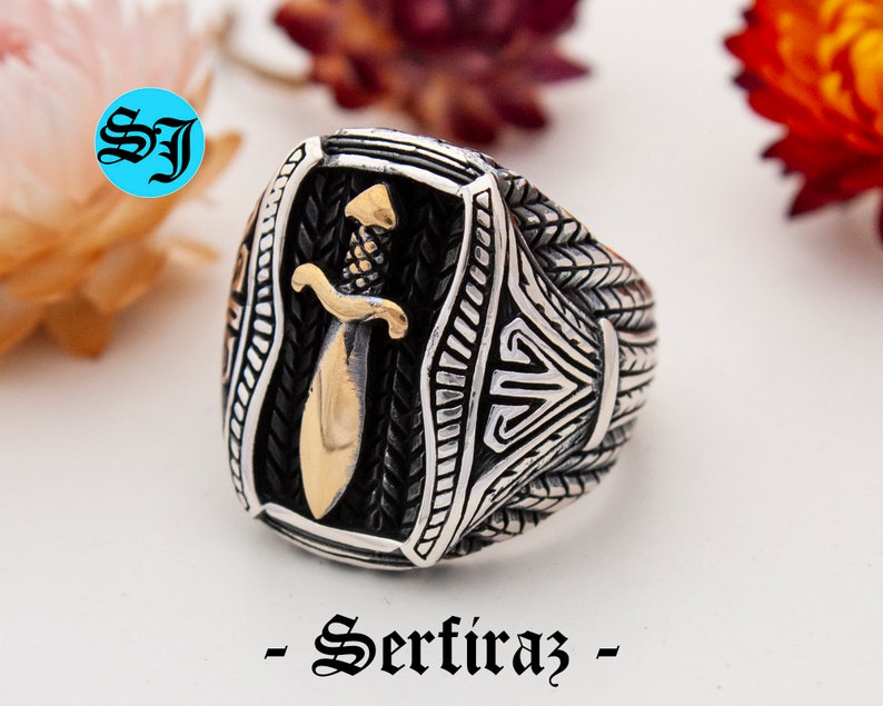 May include: A silver-toned ring with a gold-colored dagger design. The ring has intricate detailing and the text "- Serfiraz -" is at the bottom. The ring is on a white surface.