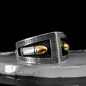 May include: A silver ring with a gold bullet design and a Greek key border. The ring is on a white stone surface.