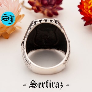 May include: Close-up of a silver ring with a black interior. The ring has a detailed, patterned design on the band. The word "- Serfiraz -" is printed in black script at the bottom of the image. The initials "SJ" are in a blue circle in the upper left corner.