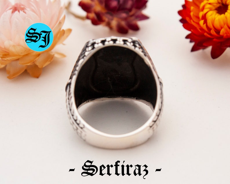 May include: Close-up of a silver ring with a black interior. The ring has a detailed, patterned design on the band. The word "- Serfiraz -" is printed in black script at the bottom of the image. The initials "SJ" are in a blue circle in the upper left corner.