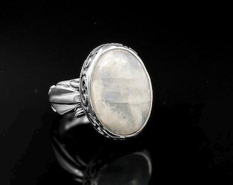 May include: A silver ring featuring an oval, iridescent moonstone. The stone is set in a detailed, ornate bezel. The ring's band has a decorative design on the sides. The ring is displayed against a black background.
