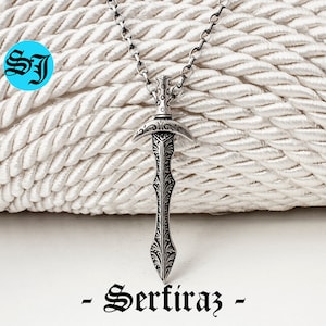 May include: A silver sword pendant necklace with intricate details and a black chain. The pendant is shaped like a sword with a pointed tip and a curved handle. The necklace is hanging on a white rope background. -Sertiraz-