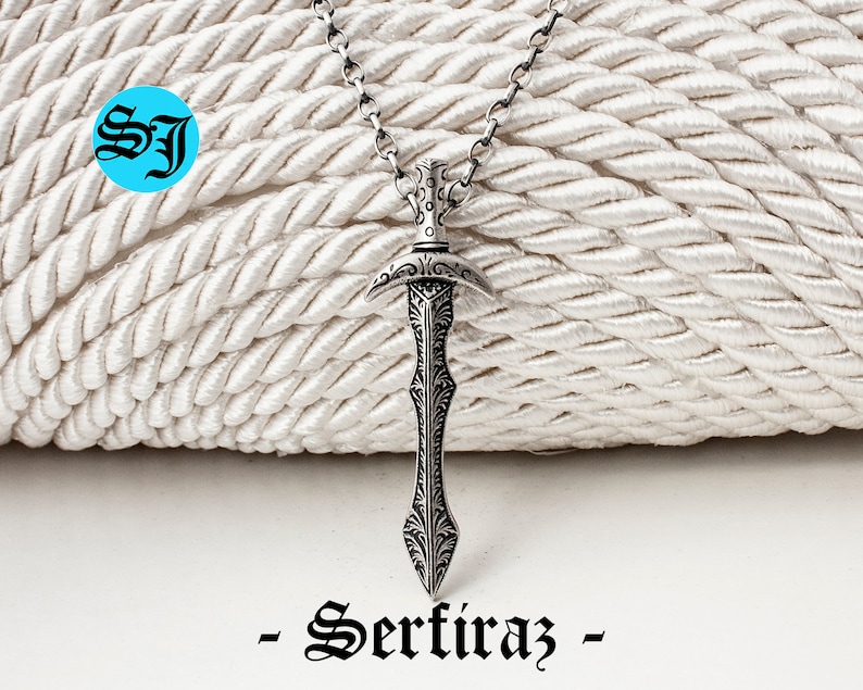 May include: A silver sword pendant necklace with intricate details and a black chain. The pendant is shaped like a sword with a pointed tip and a curved handle. The necklace is hanging on a white rope background. -Sertiraz-