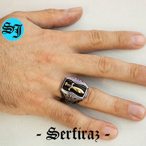 May include: A silver and gold-toned ring with a sword design. The ring has a black background with a gold sword in the center. The ring is on a hand. The text "- Serfiraz -" is at the bottom of the image.