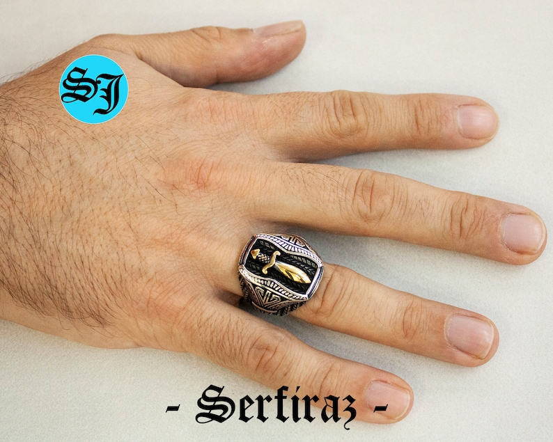 May include: A silver and gold-toned ring with a sword design. The ring has a black background with a gold sword in the center. The ring is on a hand. The text "- Serfiraz -" is at the bottom of the image.