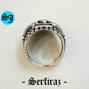 May include: A silver ring with a detailed design, featuring a central square element with a gold-colored accent. The ring has intricate patterns on the sides and the text "- Serfiraz -" below it. The initials "SJ" are in a blue circle.
