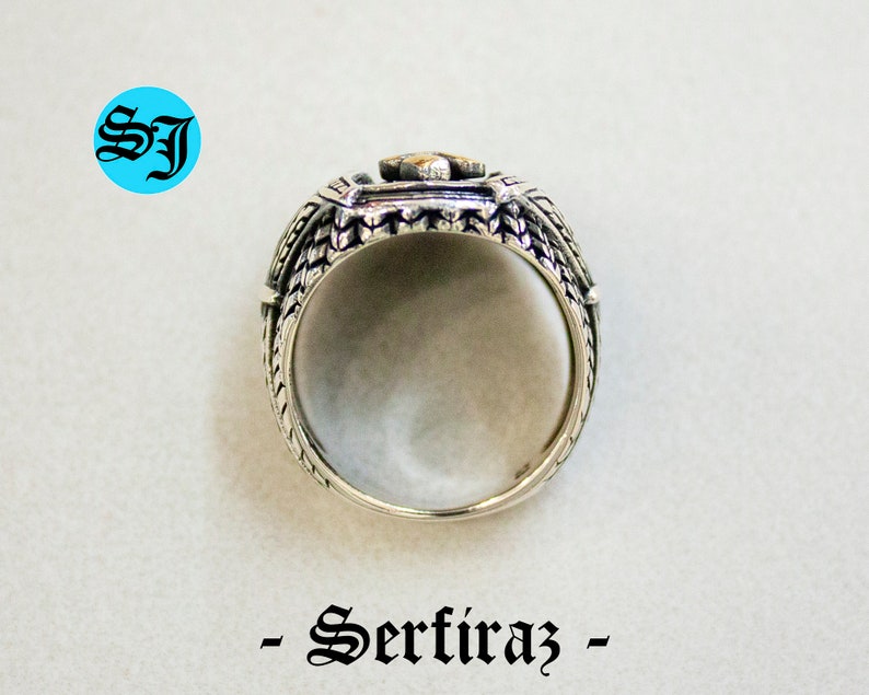 May include: A silver ring with a detailed design, featuring a central square element with a gold-colored accent. The ring has intricate patterns on the sides and the text "- Serfiraz -" below it. The initials "SJ" are in a blue circle.