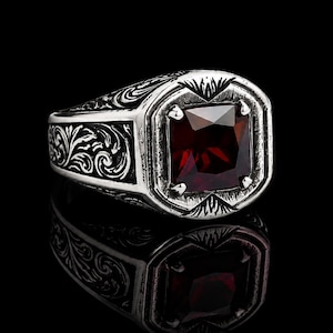 Op de afbeelding: Een zilveren ring met een vierkant geslepen dieprode edelsteen in een klauwzetting. De ringband is versierd met gedetailleerde, donker geoxideerde krulmotieven die langs de zijkanten lopen, wat zorgt voor een contrastrijke textuur.