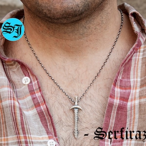 May include: A silver chain necklace with a silver sword pendant. The pendant has intricate details and a pointed tip. The necklace is worn by a person with a plaid shirt.
