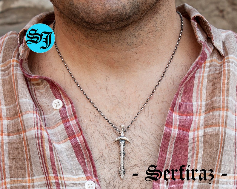 May include: A silver chain necklace with a silver sword pendant. The pendant has intricate details and a pointed tip. The necklace is worn by a person with a plaid shirt.